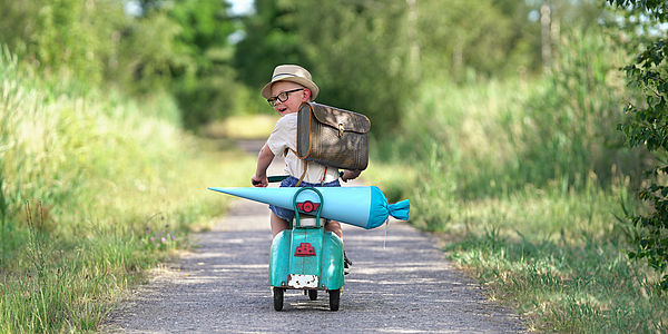 Ein kleiner Bub fährt auf einem kleinen Motorrad auf einer Straße im Grünen und blickt zurück. Er hat eine Schultasche auf dem Rücken und eine große Schultüte auf dem Gepäcksträger