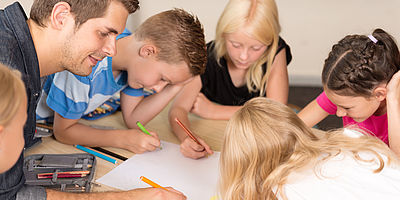 Ein Mann und Kinder sind über einen Tisch gebeugt und zeichnen gemeinsam mit bunten Stiften